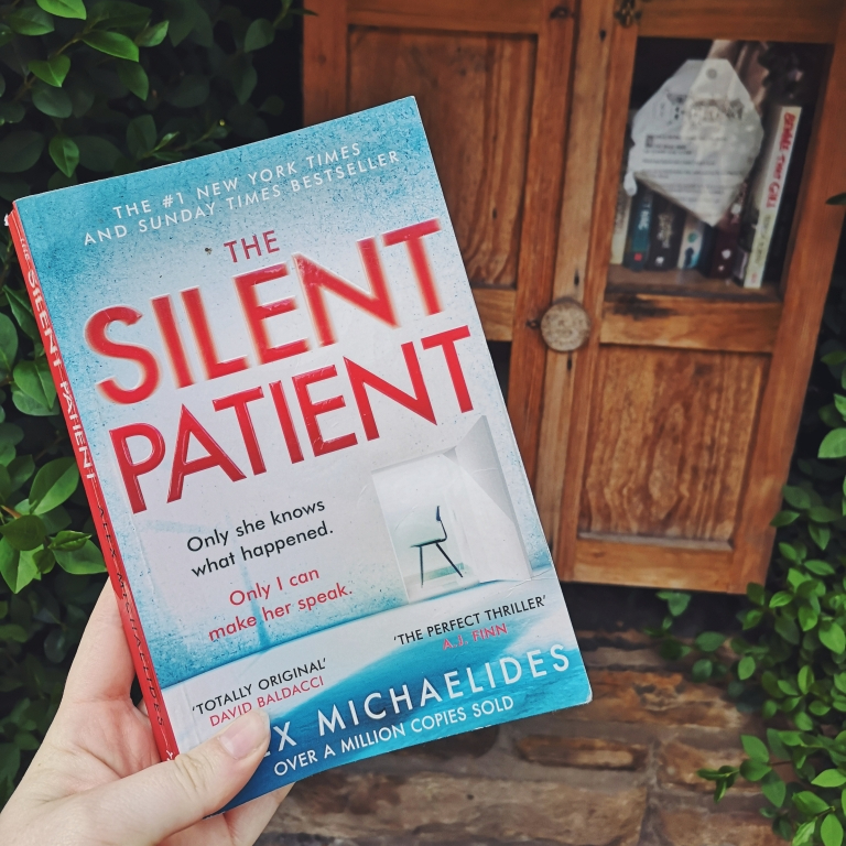 A hand holds the book The Silent Patient by Alex Michaelides out in front of a wooden shelf nestled inside a hedge. 