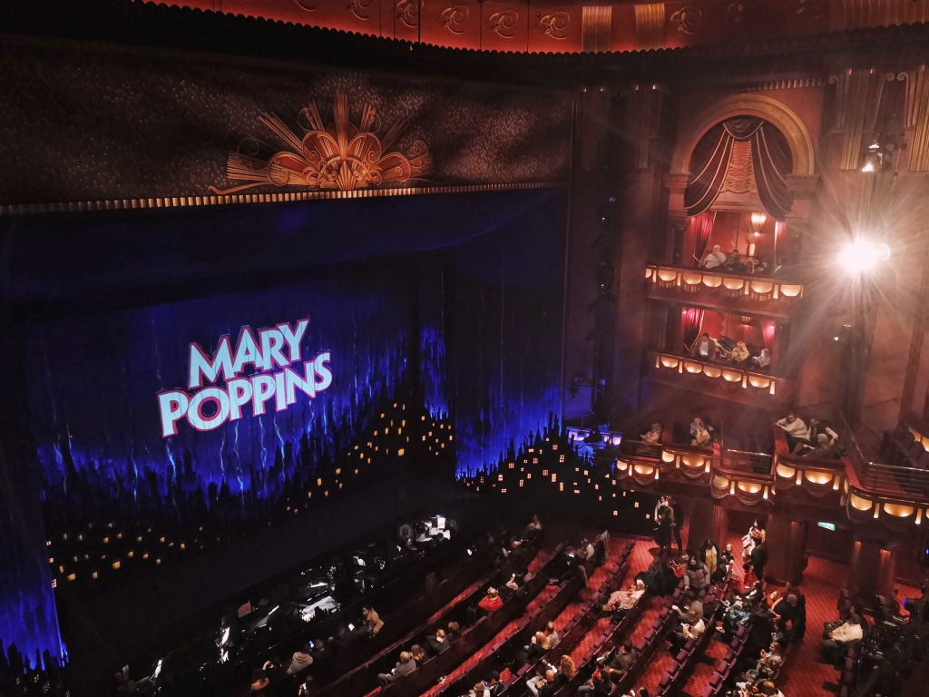 Looking down over a theatre with balconies and rows of seats. On the stage there is a sign that says Mary Poppins