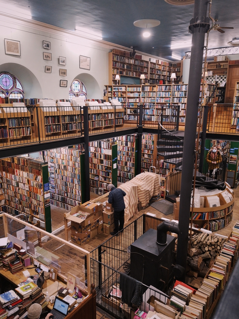 A two-floor bookshop filled with shelves and shelves full. There is a spiral staircase to the second level and a wood burning hearth in the middle of the room