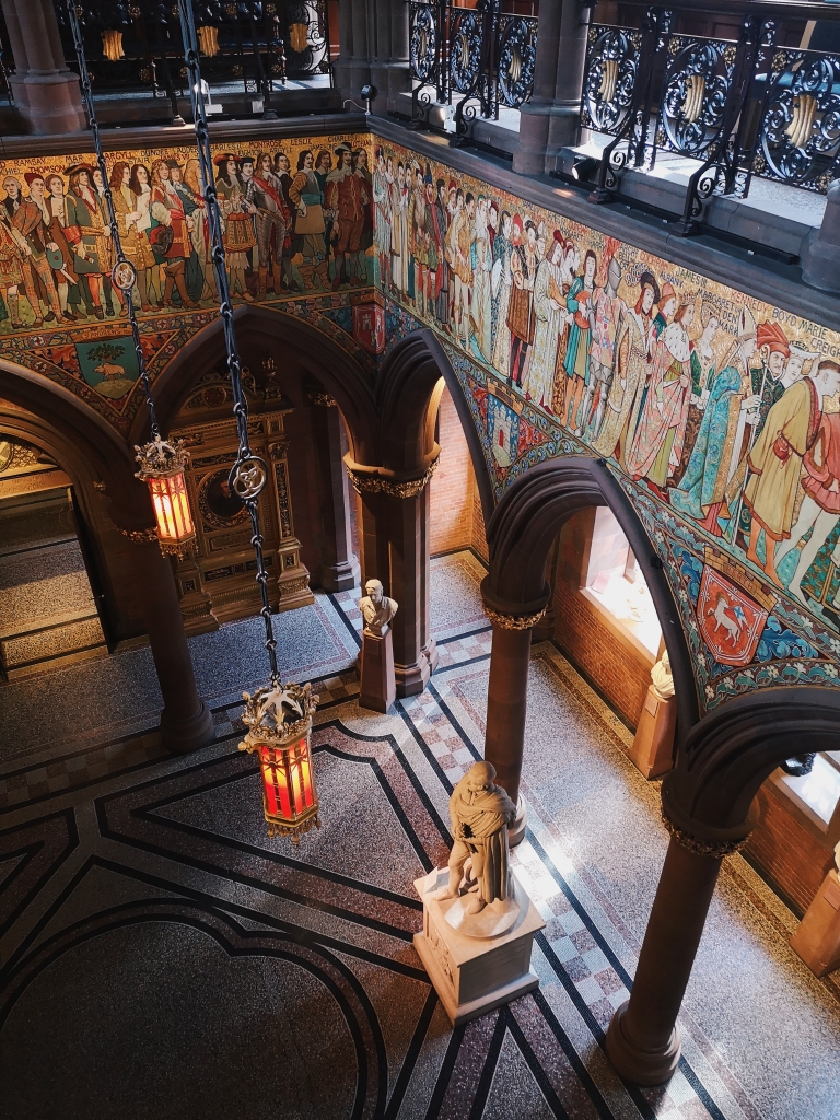 Looking down at arches and a mosaic floor. There is paintings of medieval men on the wall and a statue of Burns on the ground